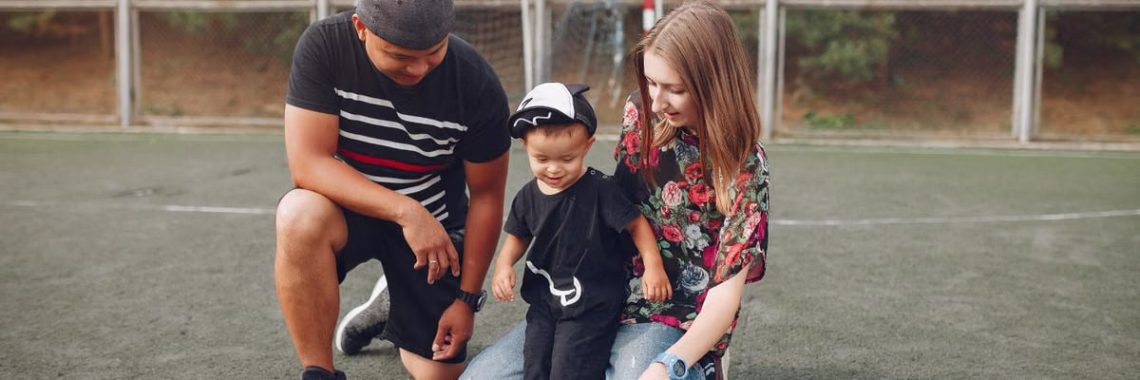 Full body of positive smiling family with cute little boy resting on football field with ball against football gate
